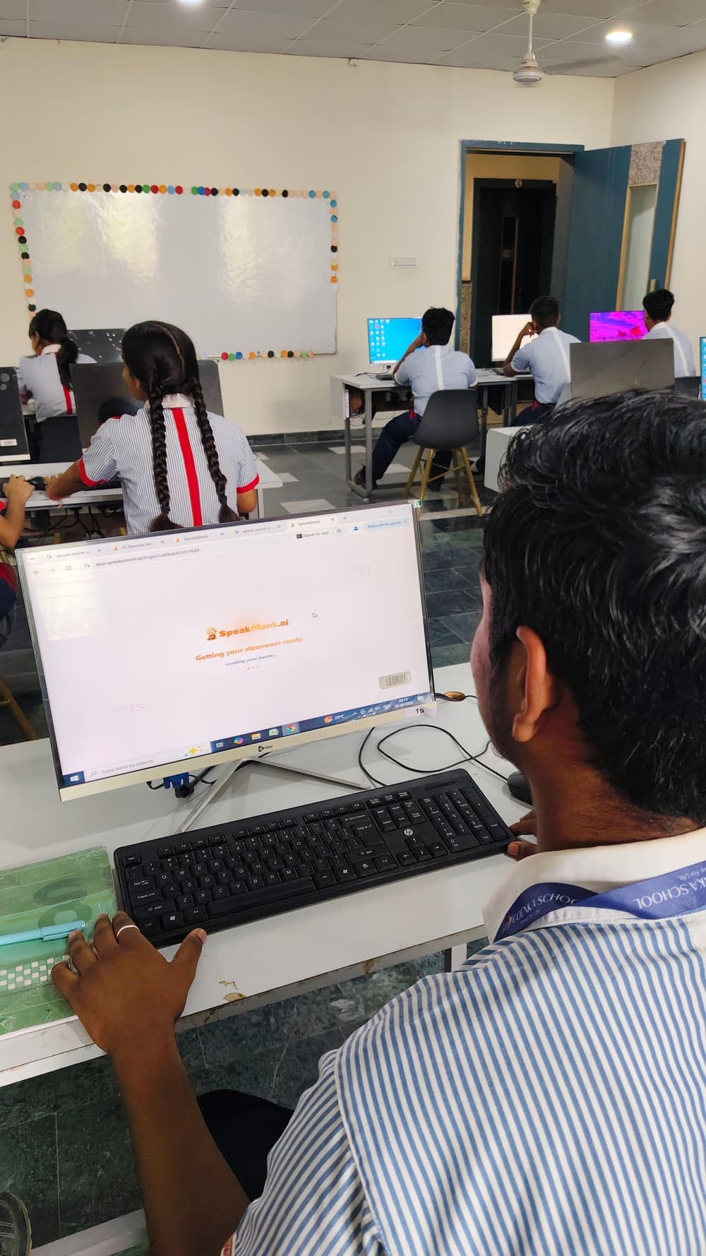 Students at desktop computers in a school lab, with SpeakMonk loading on screen